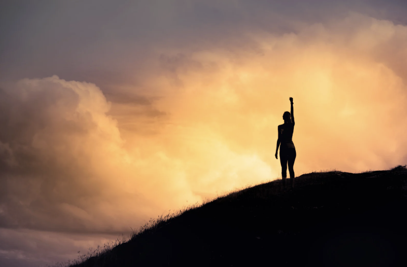 Silhouette of person standing on hill at sunset, arm raised triumphantly
