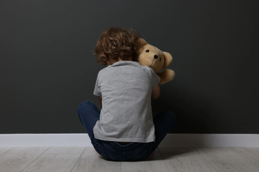 little boy sitting facing the wall holding teddy bear